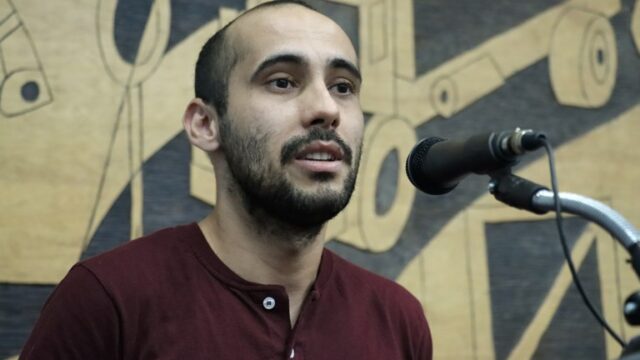 Homem com barba e camisa marsu falando ao microfone na tribuna