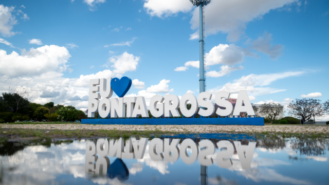Monumento com letras Eu amo Ponta Grossa e um coração azul refletido em poça d'água sob céu azul