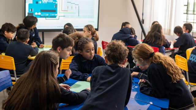 Estudantes uniformizados sentados ao redor de mesas azuis em sala de aula, atentos a uma atividade prática
