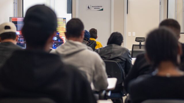 Pessoas sentadas em cadeiras acompanham apresentação em sala com placa de entrevista individual ao fundo