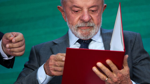 O presidente Luiz Inácio Lula da Silva de terno cinza e gravata azul com poás segura uma pasta vermelha aberta em evento e…