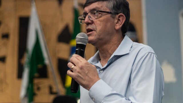 Homem com óculos e camisa social azul segura um microfone preto enquanto discursa em ambiente interno