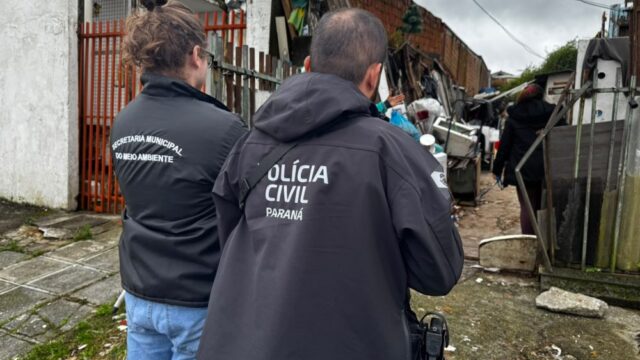 Policiais civis e agentes ambientais de costas, vestindo uniformes com identificação, em frente a uma residência