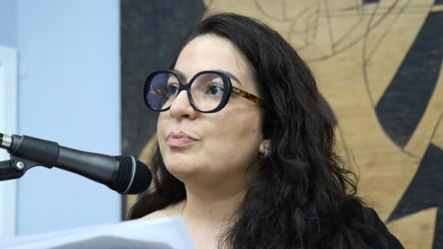 Mulher de cabelos escuros e óculos pretos fala ao microfone em tribuna