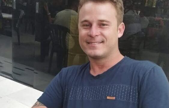 Homem de cabelo castanho claro e camisa azul sorri sentado em um estabelecimento comercial