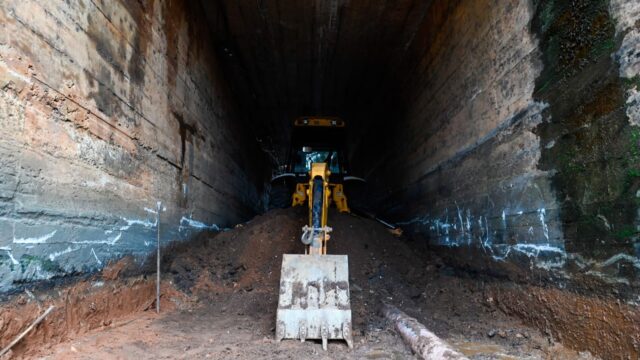 Retroescavadeira amarela operando dentro da trincheira escura com paredes de concreto e montes de terra