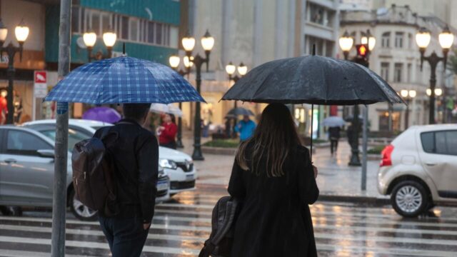 Pedestres caminham com guarda-chuvas em rua molhada durante dia de chuva em cidade paranaense