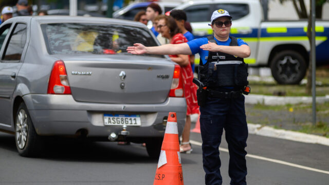 Agente de trânsito fardada orientando o tráfego próximo a um carro cinza e um cone de sinalização laranja