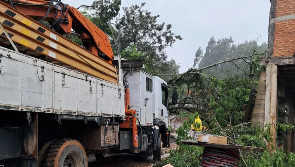 Fotografia em enquadramento médio e ângulo oblíquo de uma equipe de manutenção trabalhando após queda de árvore em área residencial. À esquerda, em primeiro plano, destaca-se a…