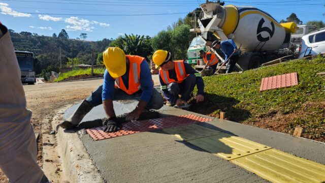 Obreiros com capacetes amarelos e coletes laranjas constroem calçada aplicando concreto e pisos táteis em Ponta Grossa