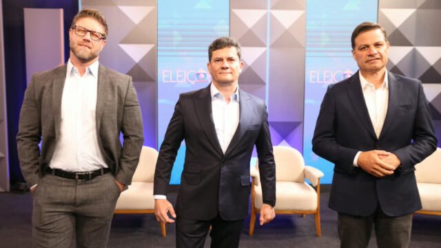 Três homens de terno posam lado a lado em estúdio de televisão