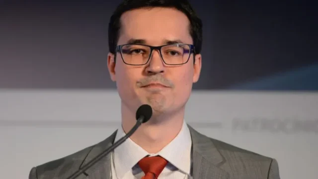 Primeiro plano de Deltan Dallagnol usando óculos, terno cinza, camisa branca e gravata vermelha diante de um microfone
