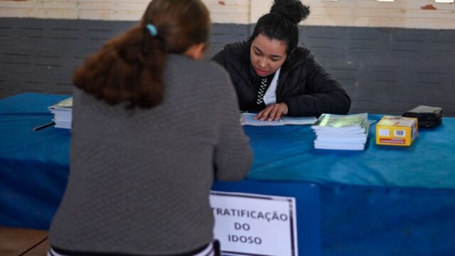 Mulher sentada de costas sendo atendida em mesa com cartaz de estratificação do idoso no bairro Chapada