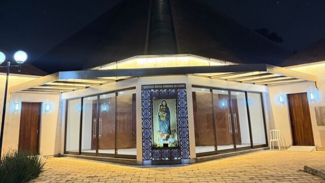 Fachada iluminada da Paróquia Nossa Senhora da Saúde à noite com mosaico religioso central