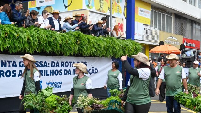 Pessoas com chapéus e roupas verdes carregam caixas de hortaliças em desfile na rua