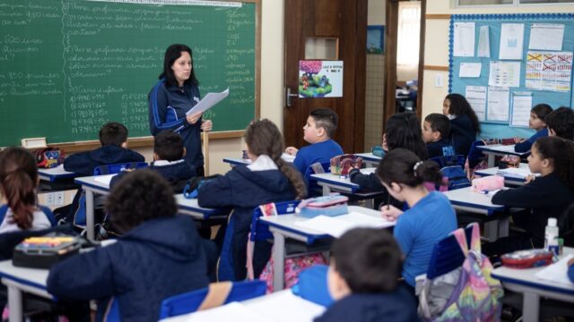 Professora em pé na frente da lousa de sala de aula com alunos atentos sentados nas carteiras