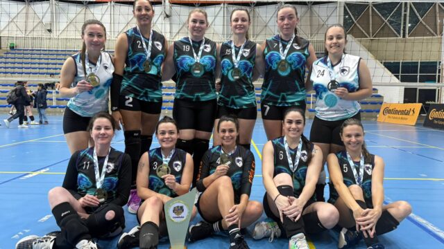 Time de voleibol feminino posa sorridente no chão e em pé com medalhas e troféu de campeão em quadra