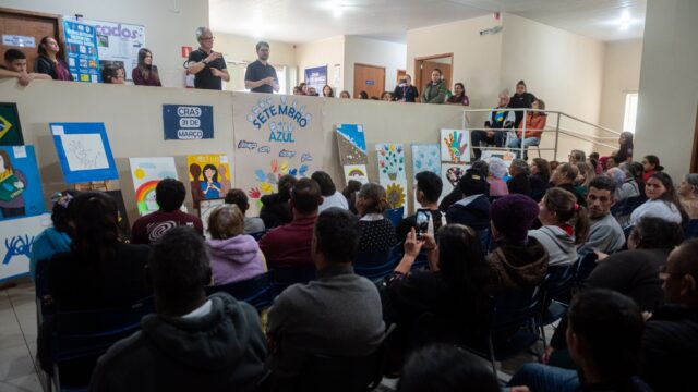 Público assiste a evento com apresentações e obras de arte em sala com painel escrito Setembro Azul