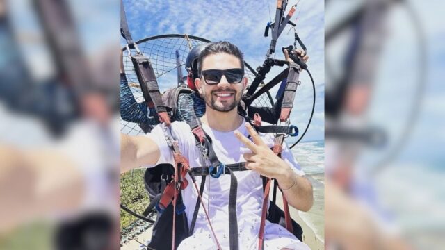 Homem sorrindo e fazendo sinal de paz e amor enquanto pilota paramotor com equipamento de segurança