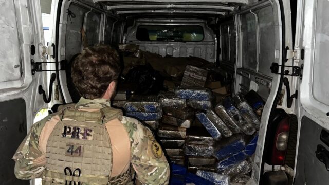 Agente da PRF fardado de costas observa carga de maconha empilhada na traseira de uma van branca aberta