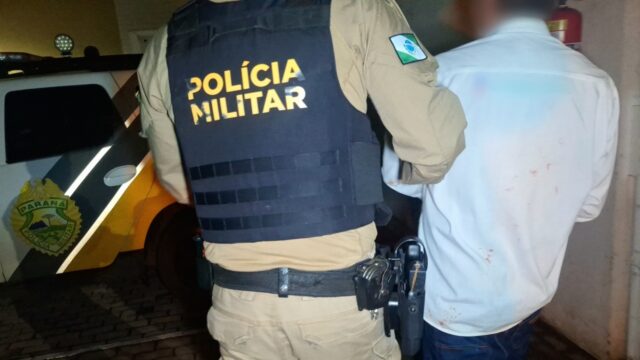 Homem algemado de costas sendo conduzido por policial militar com colete da corporação em Cascavel