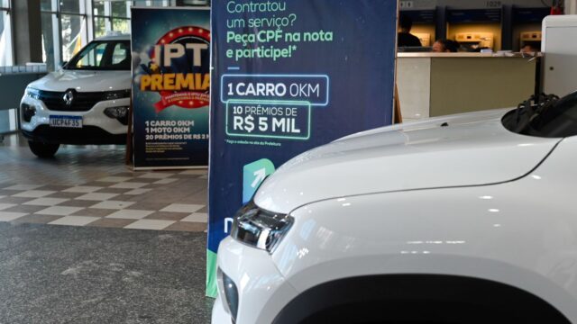 Carro branco em primeiro plano e banner azul de campanha fiscal no saguão da prefeitura