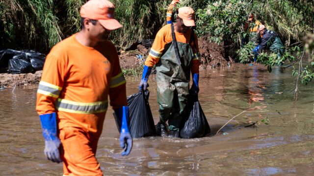 Funcionários com uniformes laranjas e botas de borracha retiram sacos de lixo de dentro de um arroio
