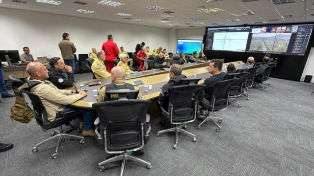 Autoridades e equipes de emergência reunidas em sala de monitoramento com painéis de dados