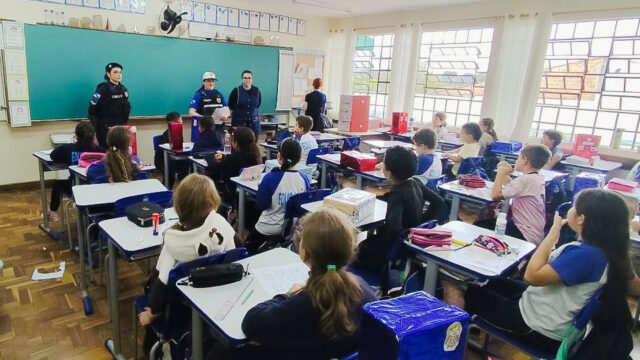 Alunos sentados em carteiras escolares em sala de aula enquanto policiais e professores conduzem atividade