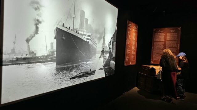 Visitantes encostados em uma parede observando uma imensa fotografia em preto e branco de um navio a vapor em uma exposição