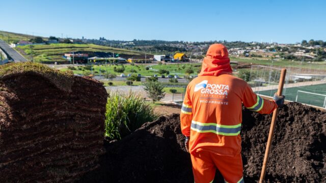 Trabalhador com uniforme laranja da Prefeitura de Ponta Grossa segurando pá perto de pilhas de terra e grama