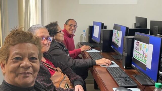 Idosos sentados em frente a computadores em sala de aula com o logotipo da FASPG na parede
