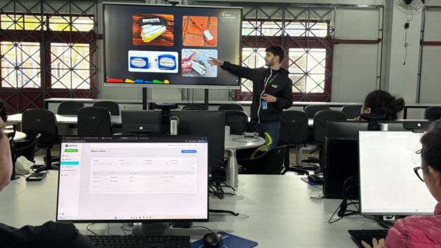 Instrutor em pé aponta para uma tela grande com fotos de baterias enquanto alunos acompanham a aula em computadores