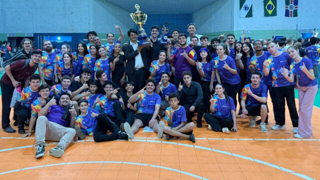 Membros da Banda Lyra dos Campos posam sorridentes com troféus em uma quadra coberta após competição no Paraná