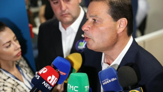 Sandro Alex falando ao microfone cercado por jornalistas e profissionais de imprensa