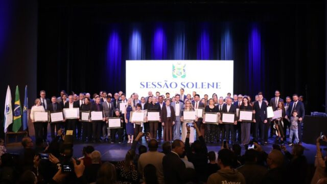 Autoridades e homenageados posam no palco com diplomas durante solenidade da Câmara Municipal