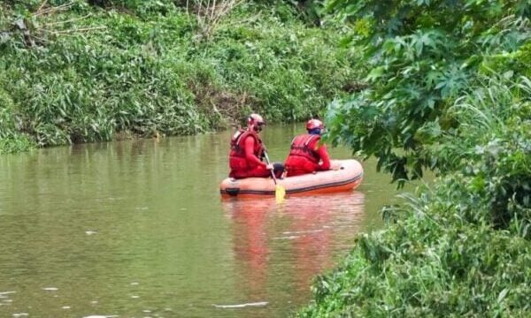 Dois bombeiros em um bote salva-vidas alaranjado navegando pelas águas de um rio cercado por vegetação densa