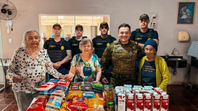 Idosas e policiais militares em farda e fardamento tático posam sorrindo atrás de uma mesa repleta de mantimentos e caixas…