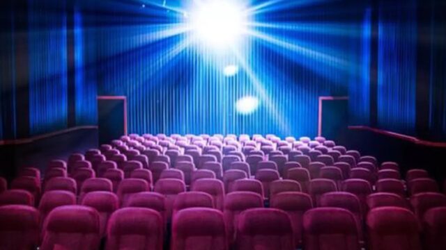 Fileira de poltronas vermelhas em sala de cinema vazia com luz de projetor acesa ao fundo