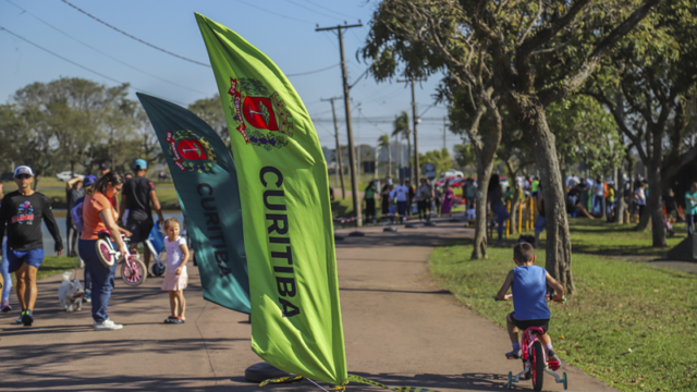 Pessoas praticam atividades de esporte e lazer em parque arborizado de Curitiba com bandeiras da cidade ao fundo