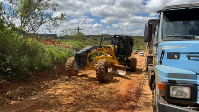 Trator motoniveladora trabalhando em trecho de estrada de terra com caminhão azul estacionado ao lado