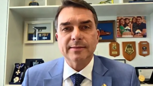 Senador Flávio Bolsonaro em videochamada com fundo de prateleiras contendo porta-retratos e placas