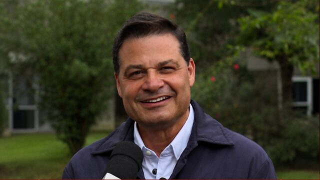 O candidato Sandro Alex sorri enquanto segura um microfone durante entrevista externa
