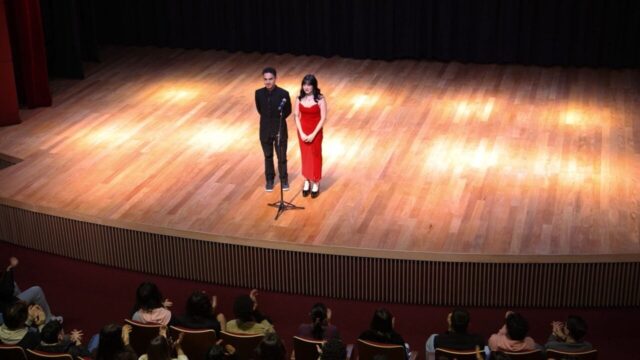 Homem de preto e mulher de vestido vermelho no palco de um auditório iluminado