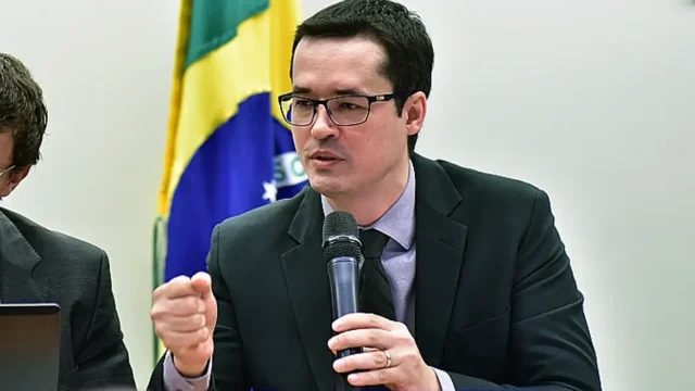 Deltan Dallagnol com terno preto e gravata falando ao microfone diante da bandeira do Brasil