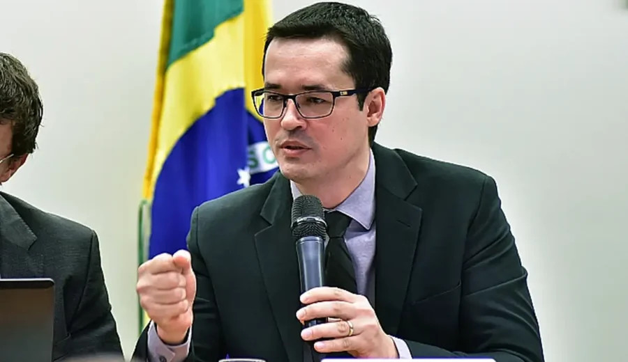 Deltan Dallagnol com terno preto e gravata falando ao microfone diante da bandeira do Brasil