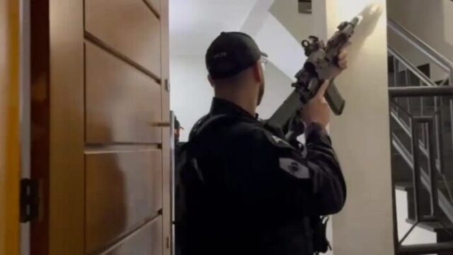 Agente de segurança com uniforme tático e fuzil realiza busca em ambiente interno de residência durante operação policial