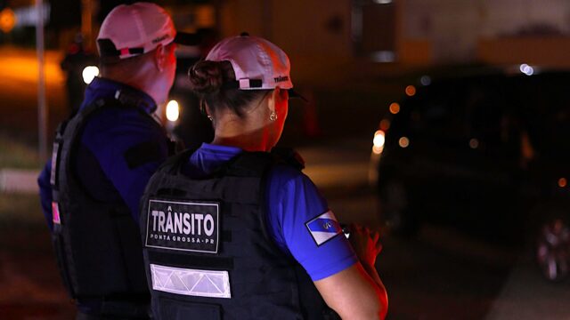 Dois agentes de trânsito fardados de costas, vestindo coletes balísticos pretos e bonés brancos durante a noite