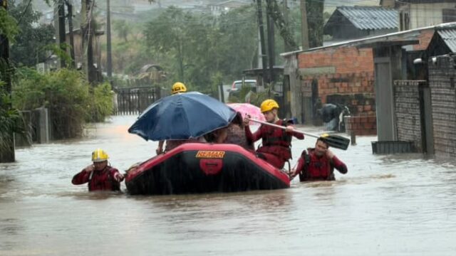 Equipe de bombeiros com capacetes amarelos em bote salva-vidas navegando por rua alagada