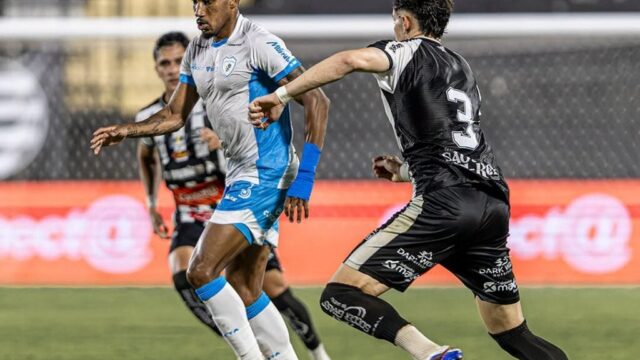Jogadores disputam a bola no gramado durante partida de futebol com placar de 2 a 2
